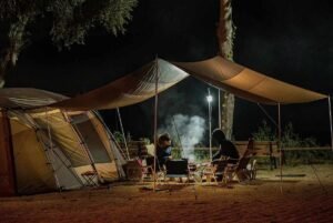 Camping nocturne avec deux personnes assises sous auvent près d'un feu de camp, entourées d'arbres