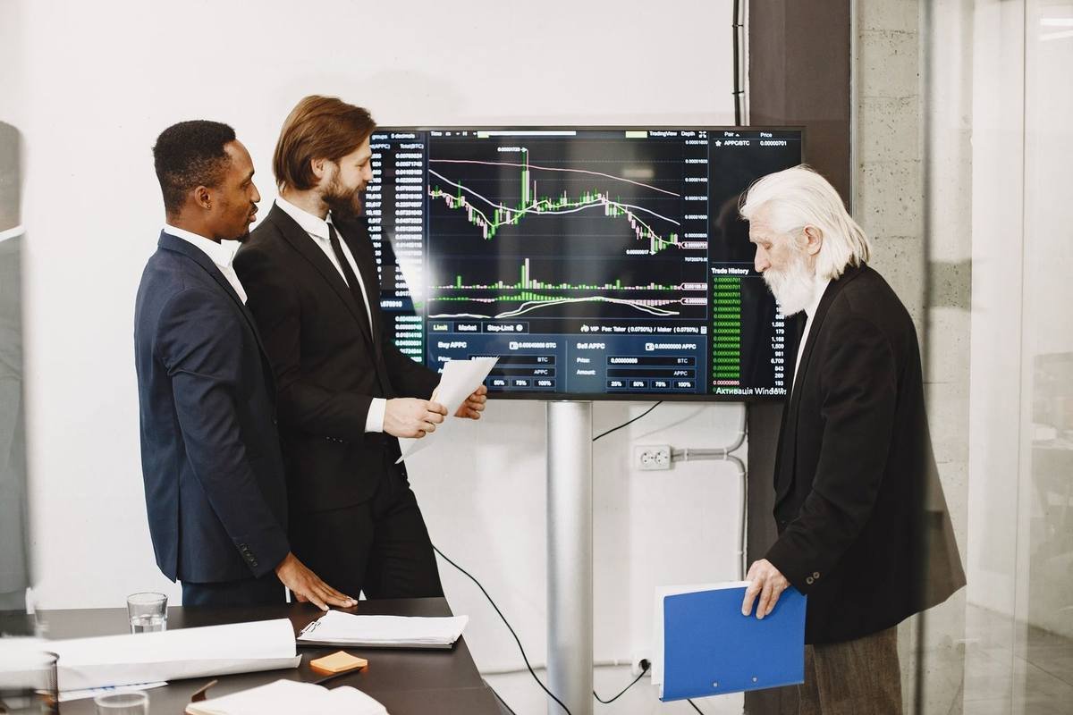 Trois hommes en réunion devant un écran de graphiques financiers, documents à la main, dans un bureau moderne