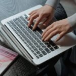Deux mains tapent sur le clavier d'un ordinateur portable blanc, assis à un bureau